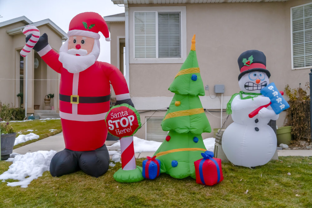 Inflatable Christmas decorations on a grassy yard with snow in winter. Santa Claus, Christmas tree, and Snowman balloons stand side by side in front of the home.