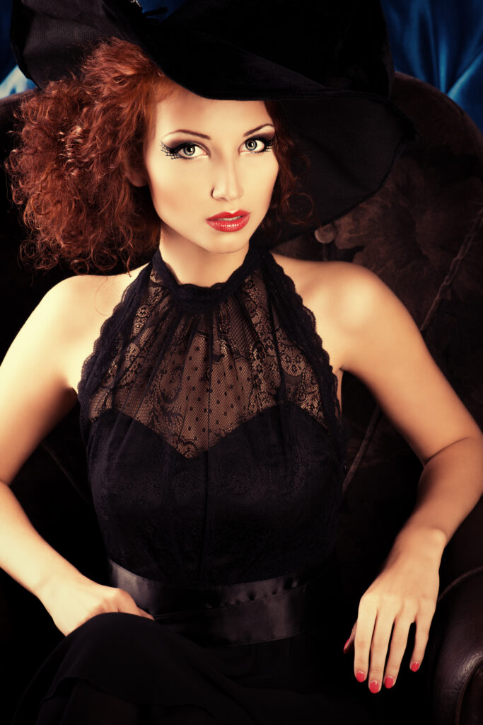 woman sitting in fancy black dress with witch hat and bright red lipstick 