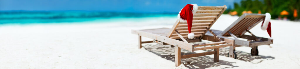 Two beach chairs on the sand with Santa hats resting on the edge. 